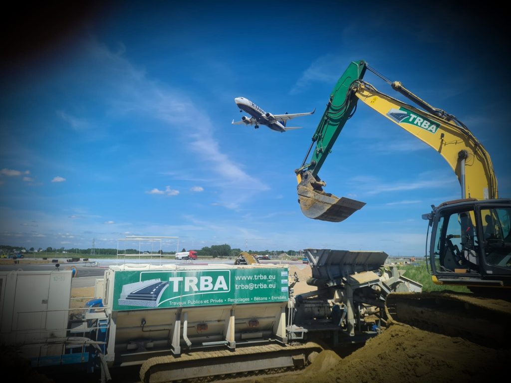 Pelle hydraulique TRBA sur le chantier de l’aéroport de Charleroi, un avion décollant en arrière-plan